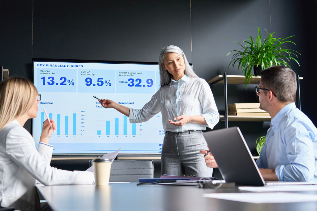 Mid age senior Asian business woman ceo executive manager showing to colleagues team income revenue data figures planning on big screen in modern office on business seminar workshop.