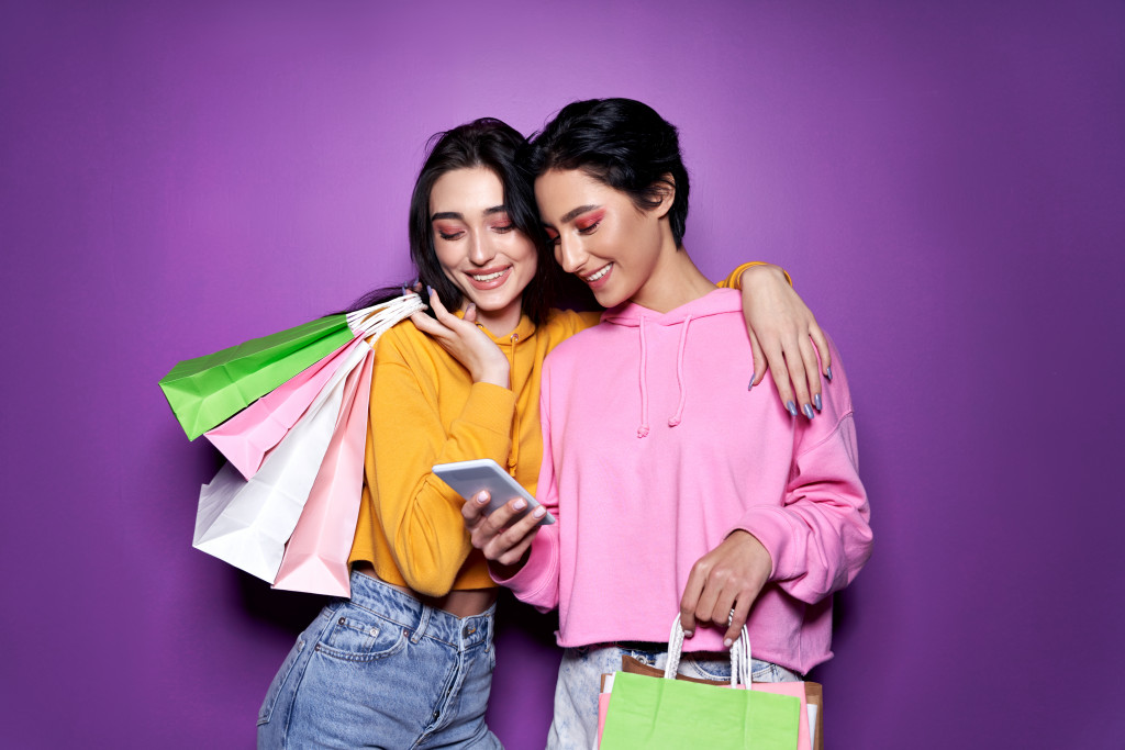 Two happy women friends shoppers holding shopping bags using mobile apps for online shopping standing on purple background.